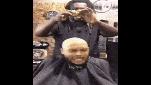 a man is getting his hair cut by a barber while wearing a cape that says fugitive .