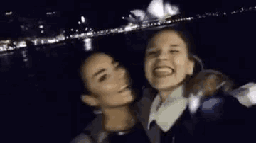 two women are posing for a selfie in front of a body of water .