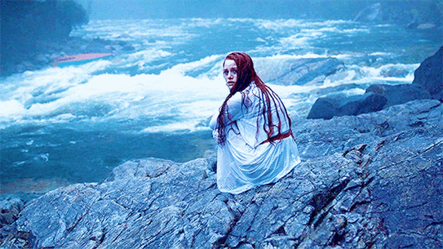 a woman with red hair sits on a rock near a river