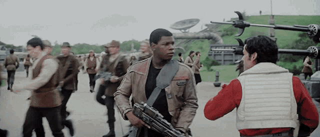 a man in a brown jacket is holding a gun and talking to another man in a red shirt