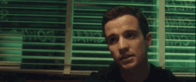 a man in a black shirt is sitting in front of a window with blinds on it