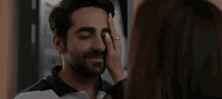 a woman is touching a man 's face while they are looking at each other .