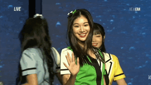 a girl in a green shirt is waving in front of a screen that says live on it