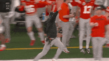 a man in a black hoodie is running on a football field surrounded by football players .