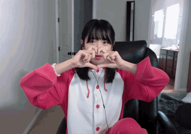 a girl in a pink and white outfit is sitting in a chair making a heart with her hands