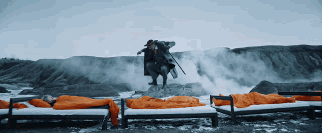 a man in a black coat is jumping over a bed with an orange sleeping bag on it