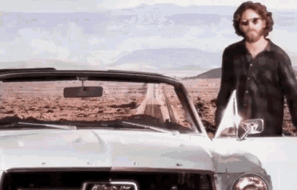 a man with a beard is standing next to a white car in the desert .
