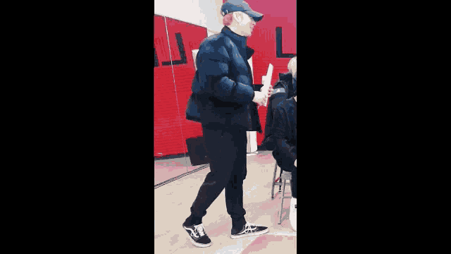 a man in a black jacket and baseball cap is standing in a room holding a piece of paper .