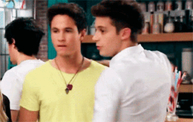 two young men are standing next to each other in a room . one of the men is wearing a yellow shirt .