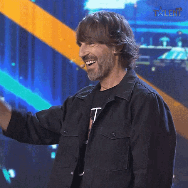 a man in a black jacket stands on a stage in front of a talent logo