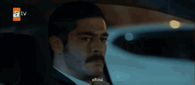 a man with a mustache is sitting in the back seat of a car with a tv logo in the background