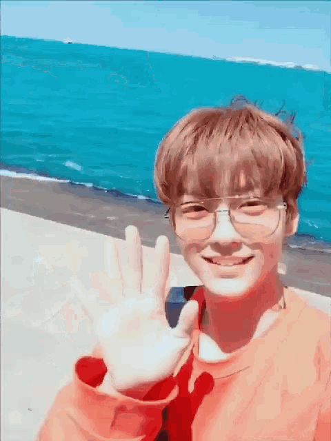 a young man wearing glasses is waving his hand in front of a body of water .