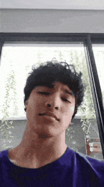 a young man in a purple shirt is making a face in front of a window .