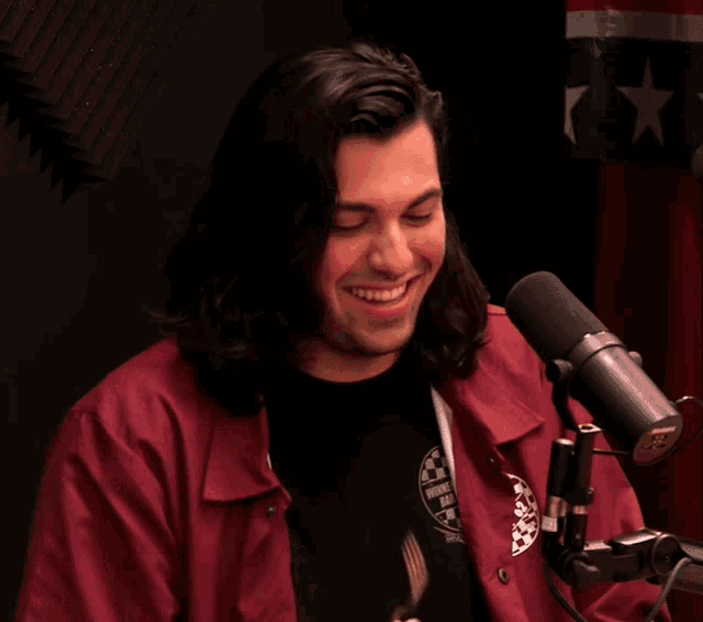 a man wearing a red jacket with a logo on the sleeve laughs in front of a mic