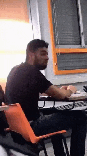 a man with a beard is sitting at a desk in a classroom in front of a window .