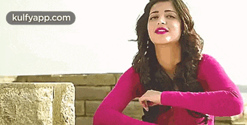a woman in a pink sweater is standing in front of a brick wall with her hands folded .