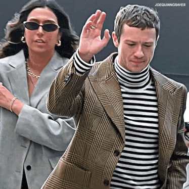 a man wearing a plaid jacket and a striped turtleneck stands next to a woman wearing sunglasses ..