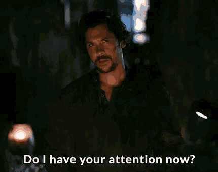 a man with a beard is standing in a dark room and saying `` do i have your attention now '' .