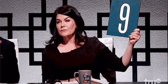 a woman is sitting at a table holding a blue sign with the number 9 on it .