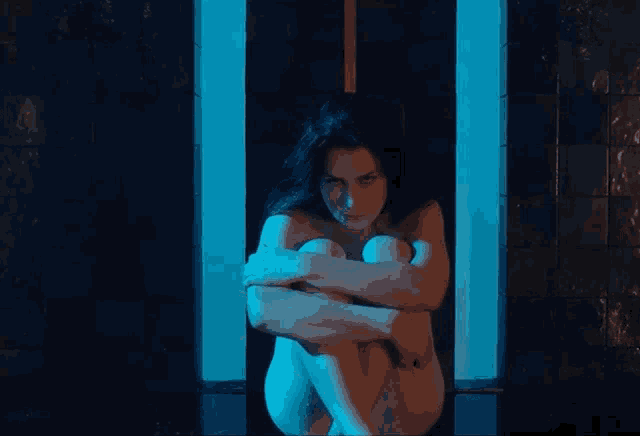 a naked woman sits with her arms around her knees in a dark room