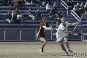 two lacrosse players on a field with jmu written on the bottom right