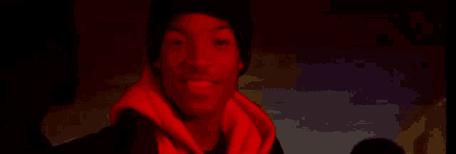 a man wearing a beanie and a hoodie is standing in a dark room with red lights behind him