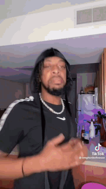 a man wearing a hooded jacket and a necklace is standing in a kitchen .