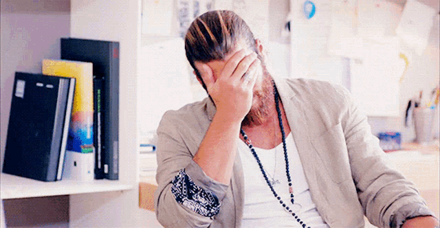 a man with a beard is covering his face with his hand in front of a bookshelf