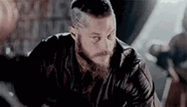 a man with a beard and a leather jacket is sitting in a room .