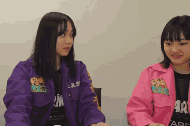 a girl wearing a purple jacket that says ' crazy world ' on it sits next to a girl wearing a pink jacket