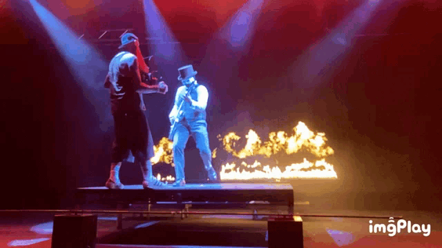 a man in a top hat stands on a stage in front of a fire behind him is a gif that says imgplay