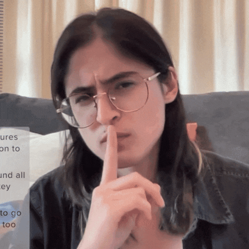a woman with long hair and glasses holds her finger to her mouth