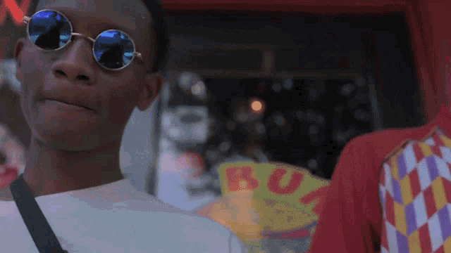 a man wearing round sunglasses stands in front of a sign that says buy