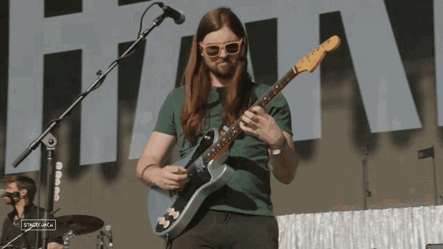 a man wearing sunglasses and a green shirt plays a guitar in front of a stage with a sign that says stagecoach