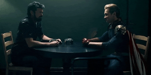 two men in superhero costumes are sitting at a table talking to each other .