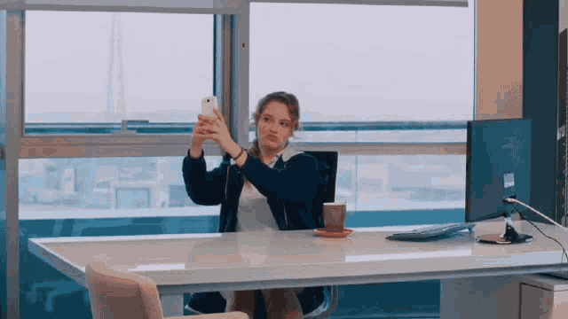 a woman sitting at a desk taking a picture of herself with her phone