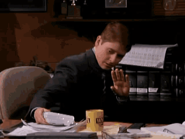 a man in a suit sits at a desk with papers and a yellow mug that says tax