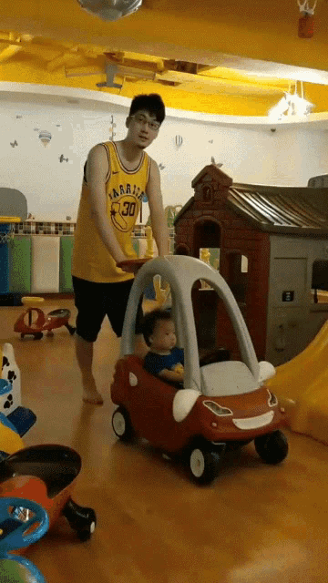 a man wearing a golden state warriors jersey is pushing a child in a toy car