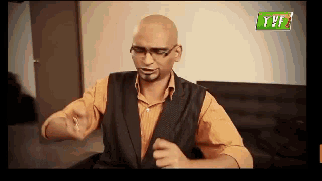 a bald man wearing glasses and a vest is sitting on a couch and making a funny face