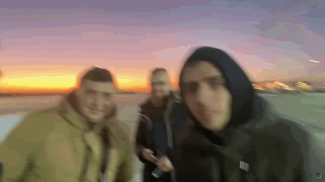 three men are standing in front of a sunset and one of them is wearing a hoodie
