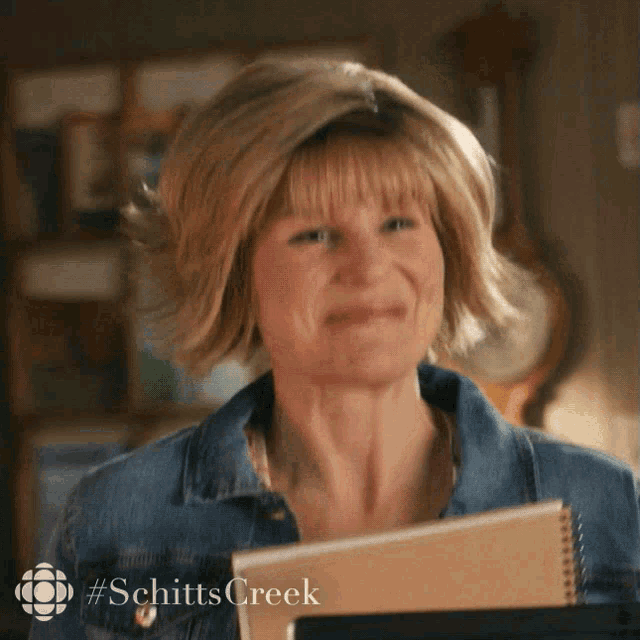 a woman in a denim jacket is smiling and holding a notebook with #schitts creek on the bottom