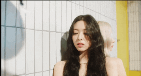 a woman with long black hair leaning against a white tiled wall