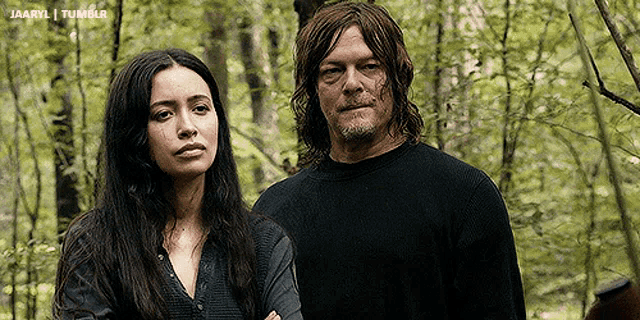 a man and a woman are standing in the woods looking at the camera .