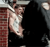 a man in a tank top leaning against a brick wall talking to another man .