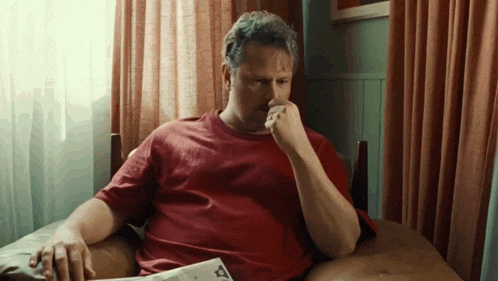 a man in a red shirt is sitting on a chair reading a newspaper
