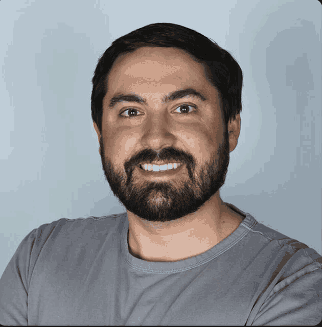 a man with a beard wears a grey shirt and smiles for the camera