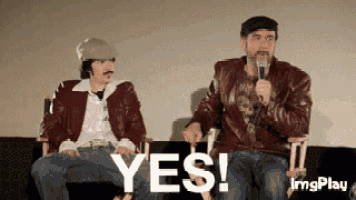 two men are sitting next to each other on a couch and one is holding a microphone and the other is saying yes .