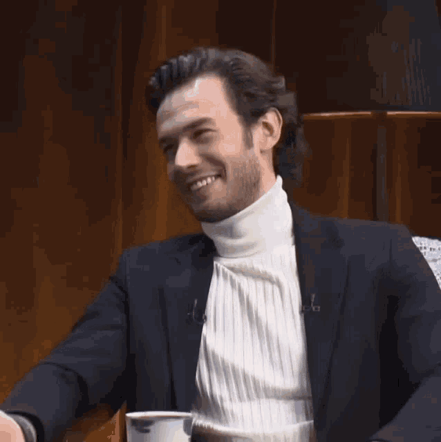 a man wearing a black jacket and a white turtleneck is sitting at a table with a cup of coffee