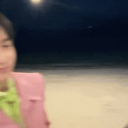 a blurry picture of a woman in a pink jacket and green shirt standing on a beach at night .