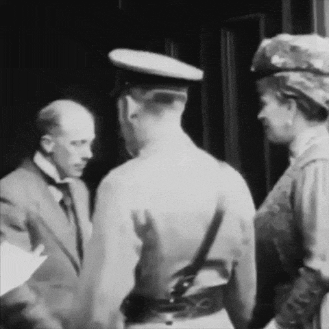a black and white photo of a man in a suit and tie talking to a woman in a hat